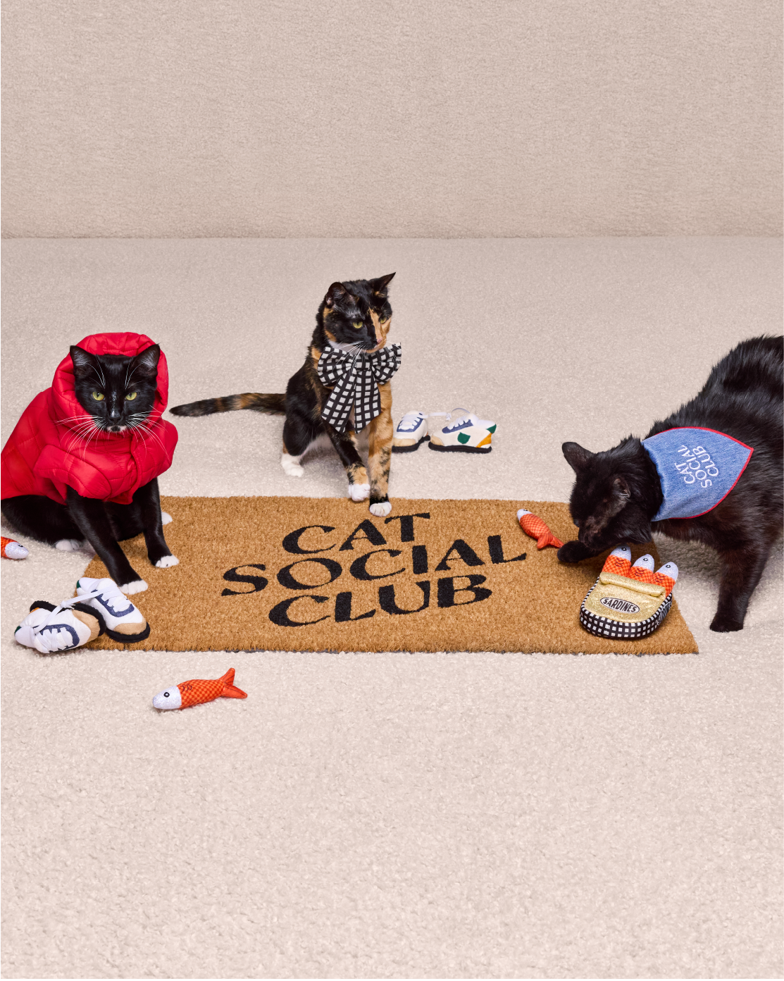 Three cats wear items from The Cuddle Collab and stand around a doormat that reads "Cat Social Club."