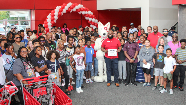 A group of people, including Bigs, Littles and Target team members, gather for a group photo inside a Target store during a back-to-school event. Bullseye, the Target mascot, stands in the center of the group under a red and white balloon arch.