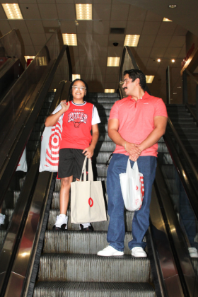 Two people, one a Big and the other a Little, ride down an escalator carrying Target shopping bags during a back-to-school event.