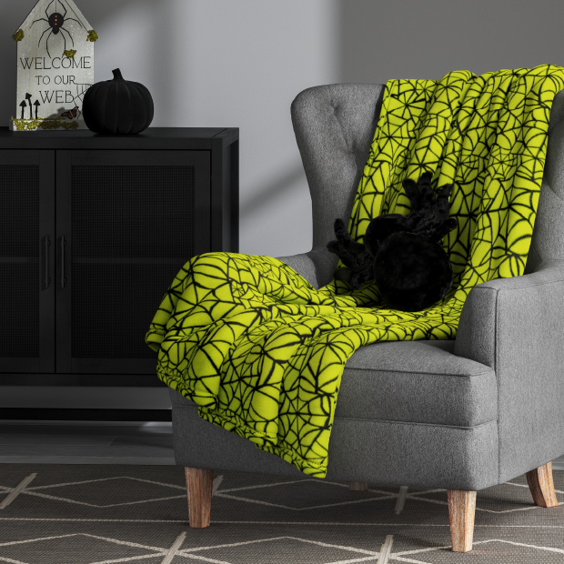 A decorative black spider-shaped throw pillow and a neon blanket with a black cobweb design laying across a gray upholstered chair, next to Halloween decor on top of a black cabinet.